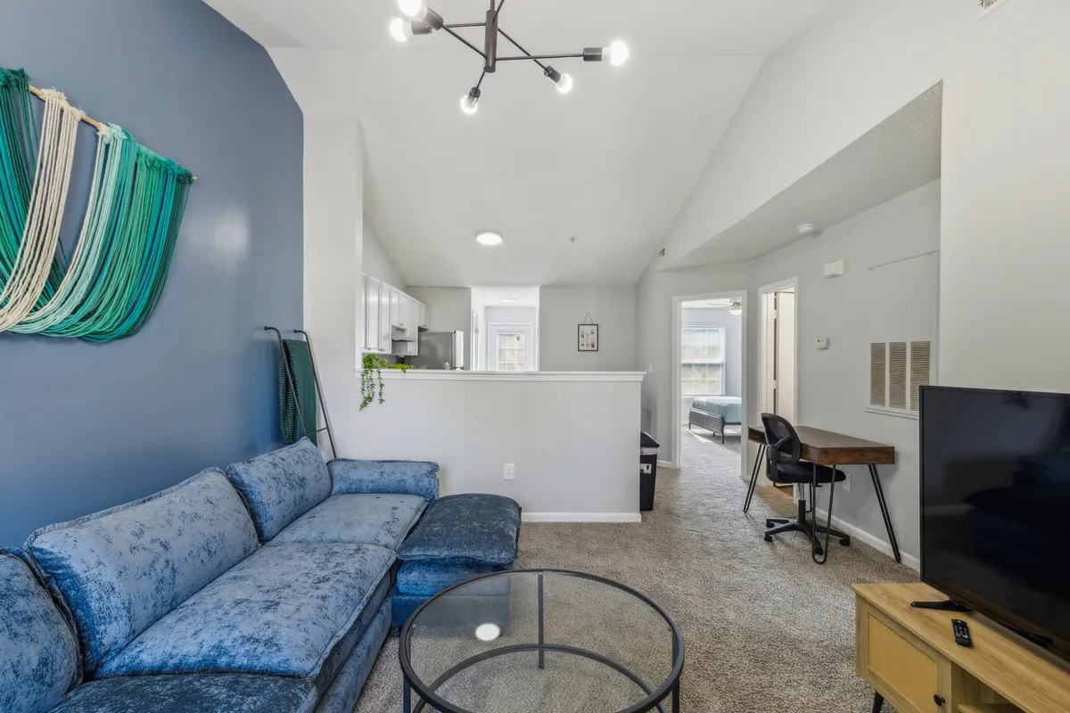 A modern living area featuring a blue couch, elegant chandelier, glass coffee table, and artistic wall decor. The room exudes a cozy and modern mood, ideal for both relaxation and entertaining.