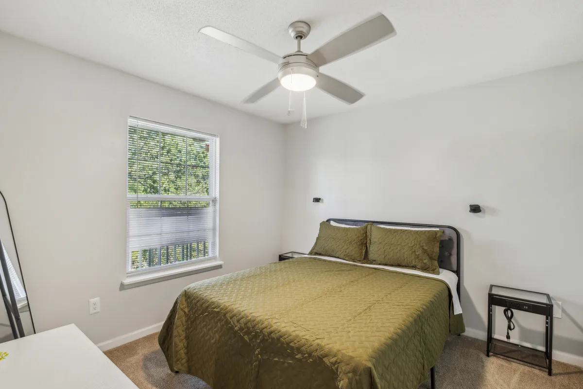 This image features a plush queen bed with inviting green bedding, a window with blinds that hint at the room's ability to block or filter light, and a ceiling fan above. The tranquil and cozy mood is palpable, with natural sunlight gently streaming through the window, suggesting a serene start or end to the day.