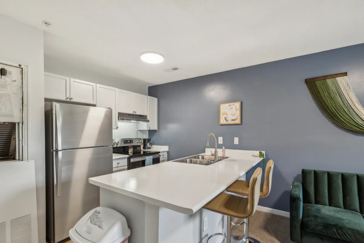 A spacious modern kitchen featuring a central island, stainless steel appliances, a dramatic dark blue accent wall, and adjoining living area with a green couch and decorative wall art. The setup conveys an inviting, practical space for cooking, dining, and socializing.