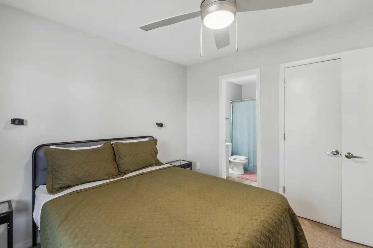 A relaxing bedroom scene showcasing the queen bed with green bedding, complemented by nightstands that add functional elegance. A doorway to the bathroom is visible, enhancing the sense of privacy and suite-like comfort. The soft lighting sets a calming atmosphere, perfect for unwinding.