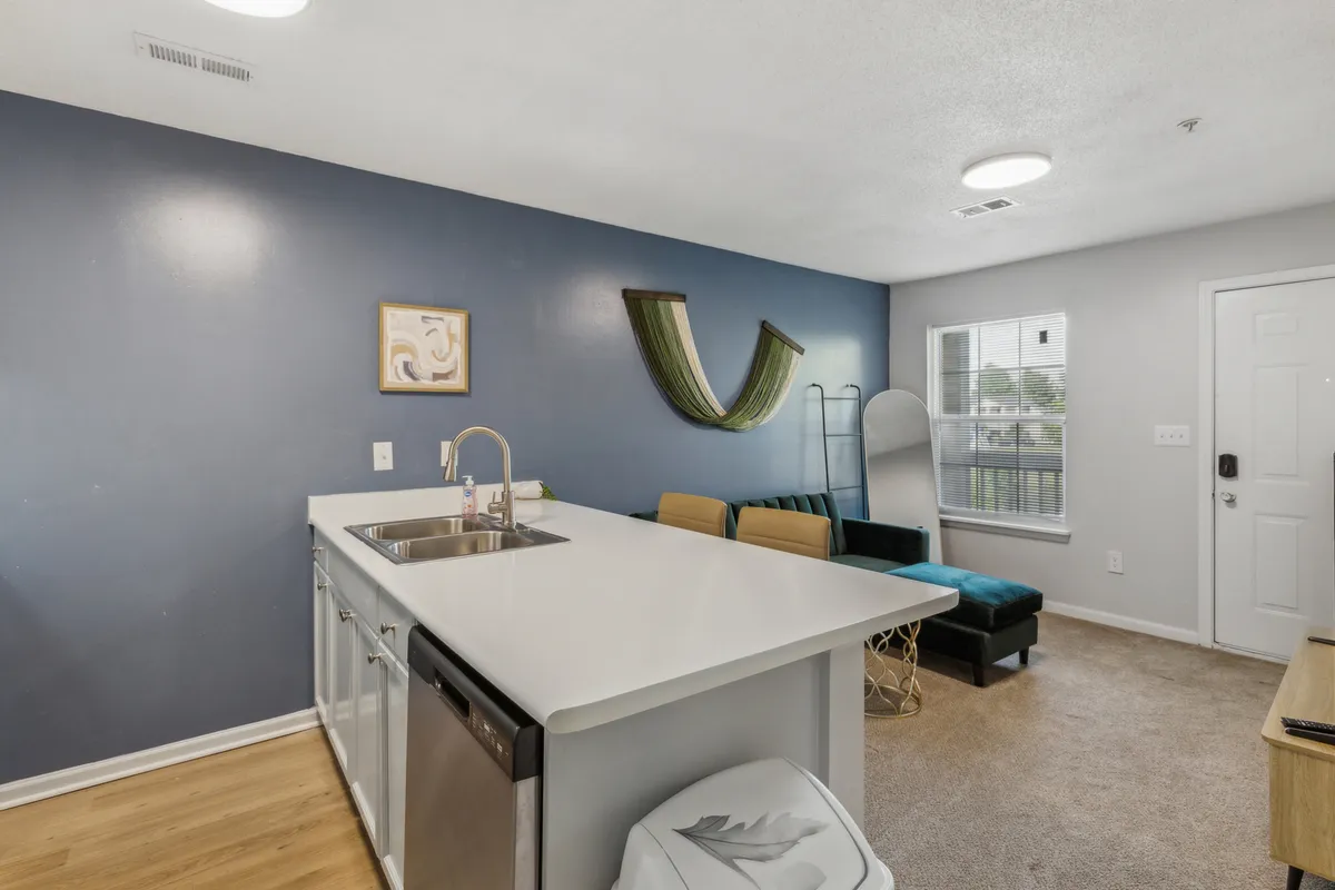 A bright, open kitchen scene with a central island, green wall art, and large window letting in natural light. Comfortable sofa seating is nearby, creating a seamless flow between kitchen, dining, and relaxation zones.