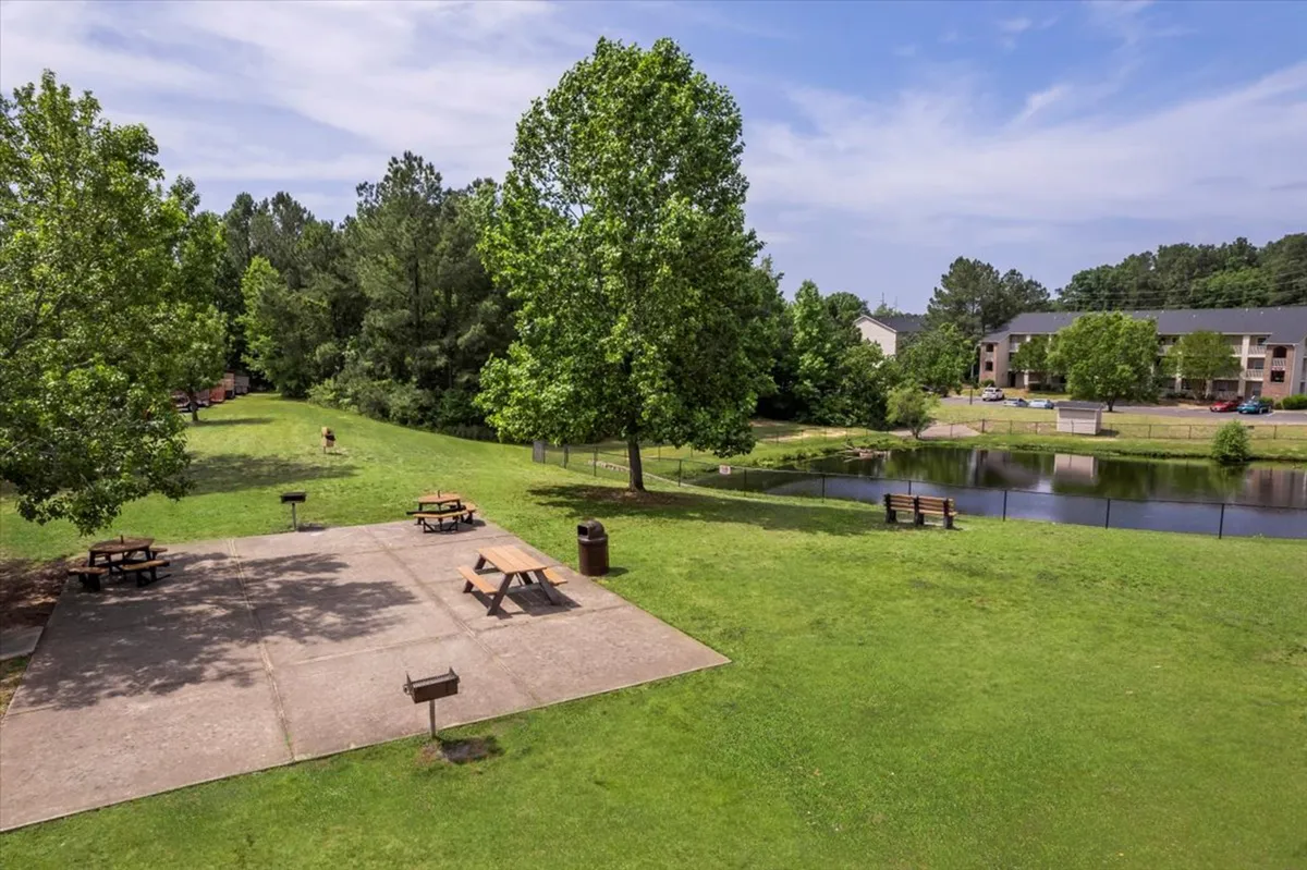 A tranquil outdoor area featuring picnic tables, a pond flanked by trees, and benches ideal for relaxing. The space is clearly designed for both quiet moments and social gatherings, with opportunities for fishing, walking, or attending community events.
