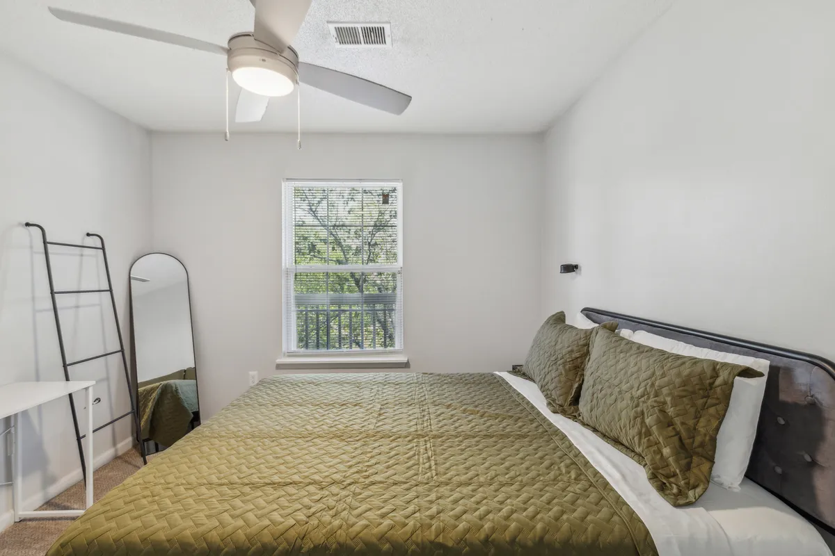A serene bedroom view focusing on the queen bed with a green quilt, a window allowing gentle natural light, a mirror, and a ceiling fan overhead. The scene feels peaceful and airy, inviting guests to rest or read in comfort.