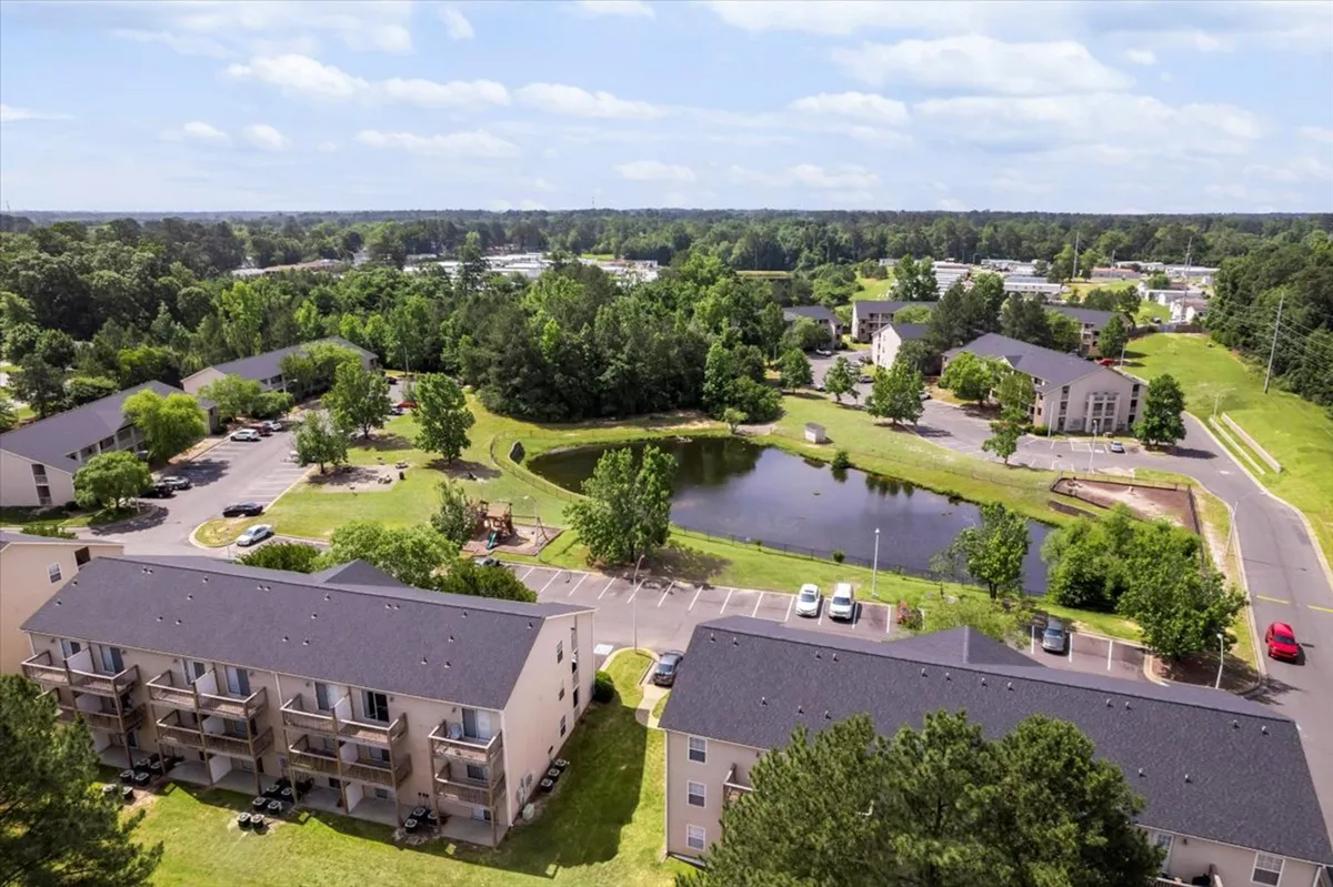 This exterior image features a serene pond surrounded by green trees, with apartment buildings and a parking area in the background. The inviting scene showcases walking paths and open green spaces suitable for picnics or a morning jog, set against the backdrop of a relaxed community environment.
