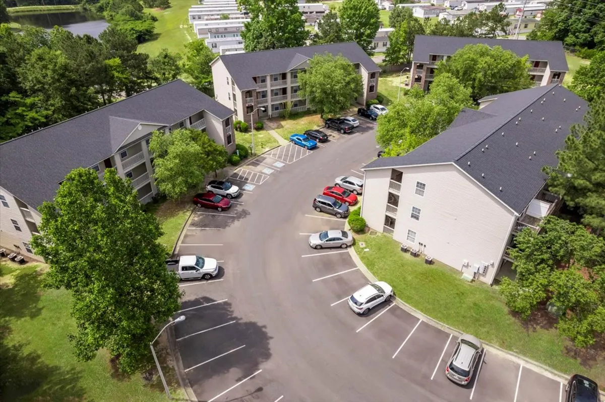 This image showcases the apartment buildings surrounded by green trees, with a visible parking lot and several cars. The scene feels inviting and peaceful, indicative of a well-maintained, accessible residential community.