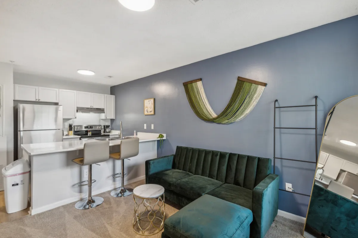 A contemporary and inviting living space with a green velvet sofa, round coffee table, kitchen island, and stylish wall decor. The area is bathed in daylight, emphasizing comfort and sociability.