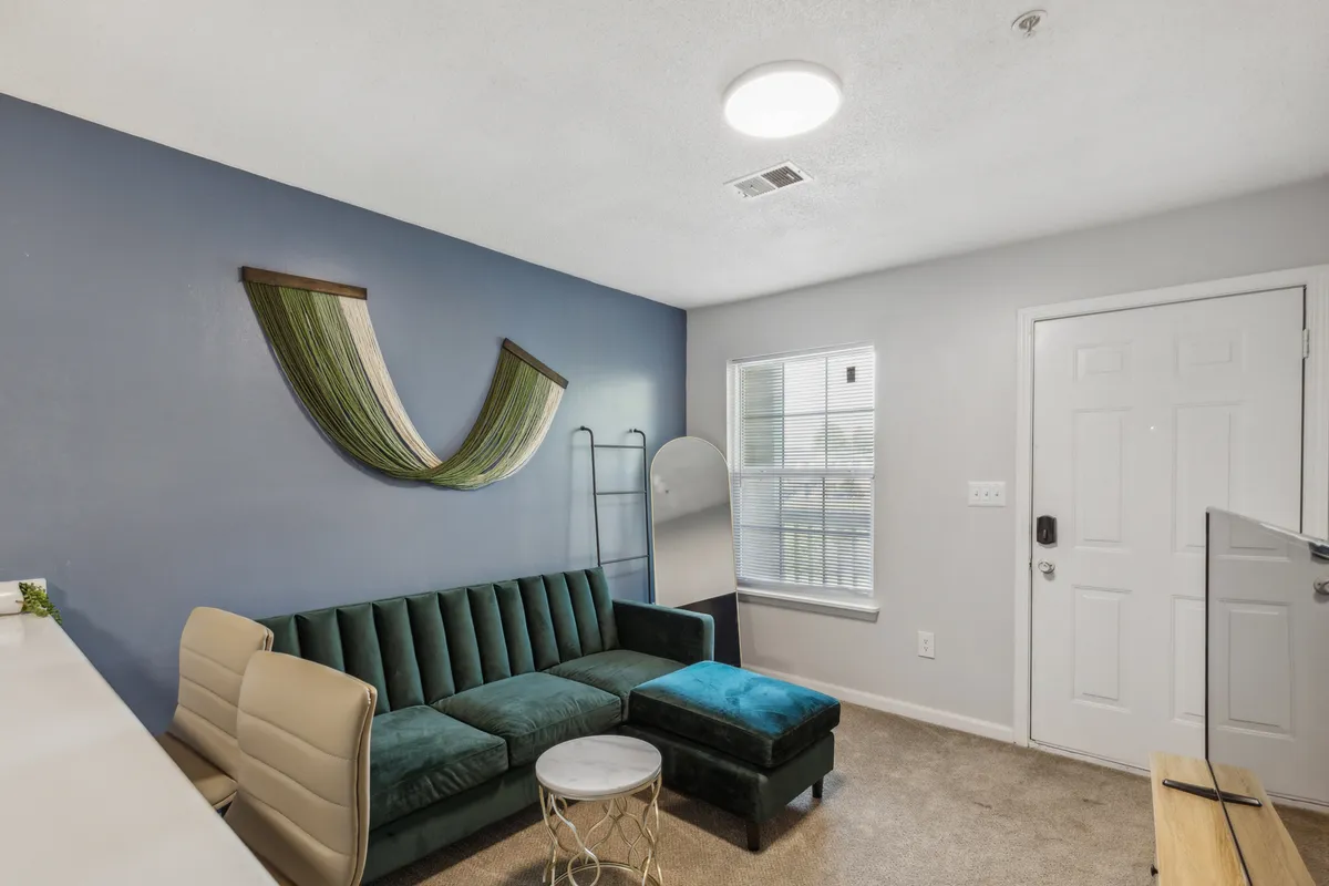 The image features a contemporary interior with a plush green velvet sofa as the centerpiece, complemented by stylish decorative wall art and a statement mirror near the entryway door. The setting radiates a cozy, inviting atmosphere ideal for relaxing, socializing, or simply unwinding after a day exploring Fayetteville.