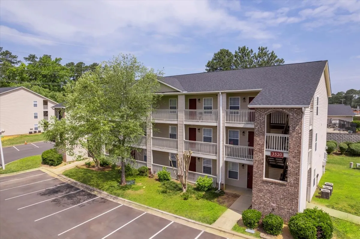 A bright exterior shot highlighting an apartment building with greenery and a parking area. The scene evokes a sense of community, with features that suggest outdoor relaxation and morning routines on balconies.